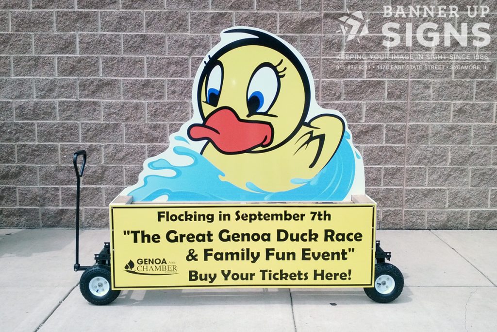A custom cut corrugated sign, shaped like a duck, attached to a small wagon advertises an event.