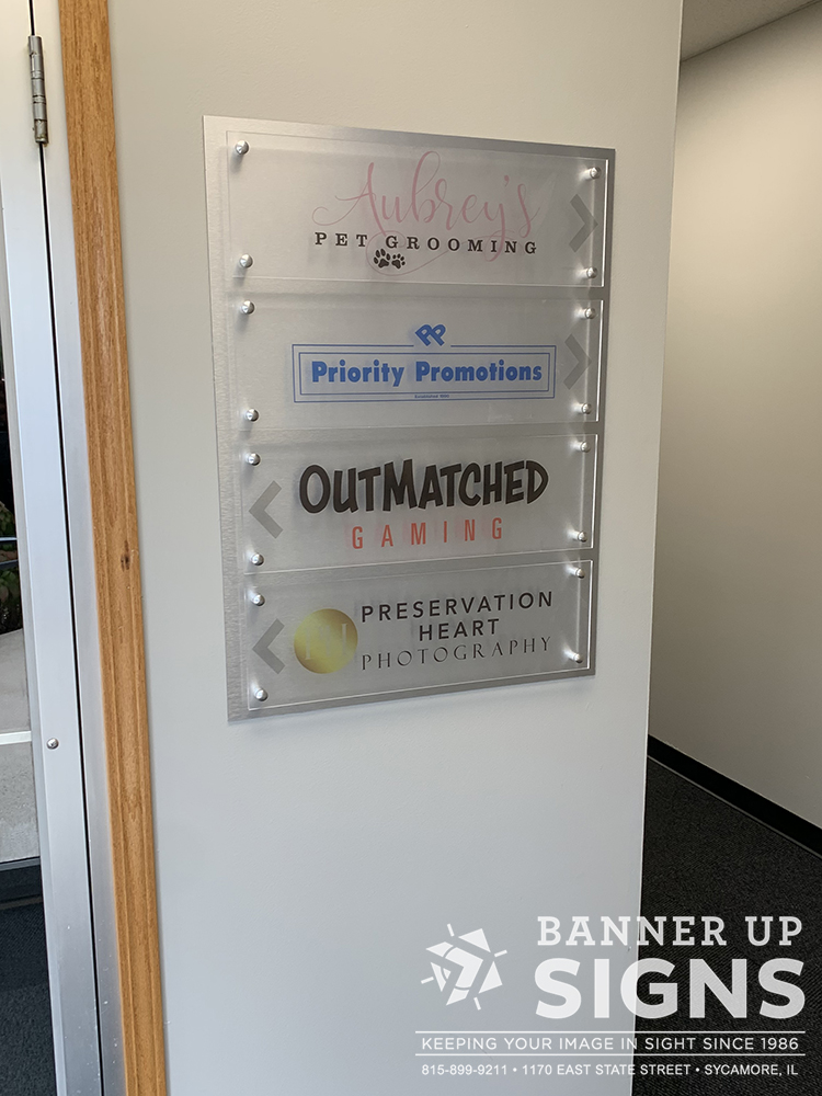 A brushed aluminum backer with 4 acrylic panels are used as a directory of businesses and which direction to go to find their location.