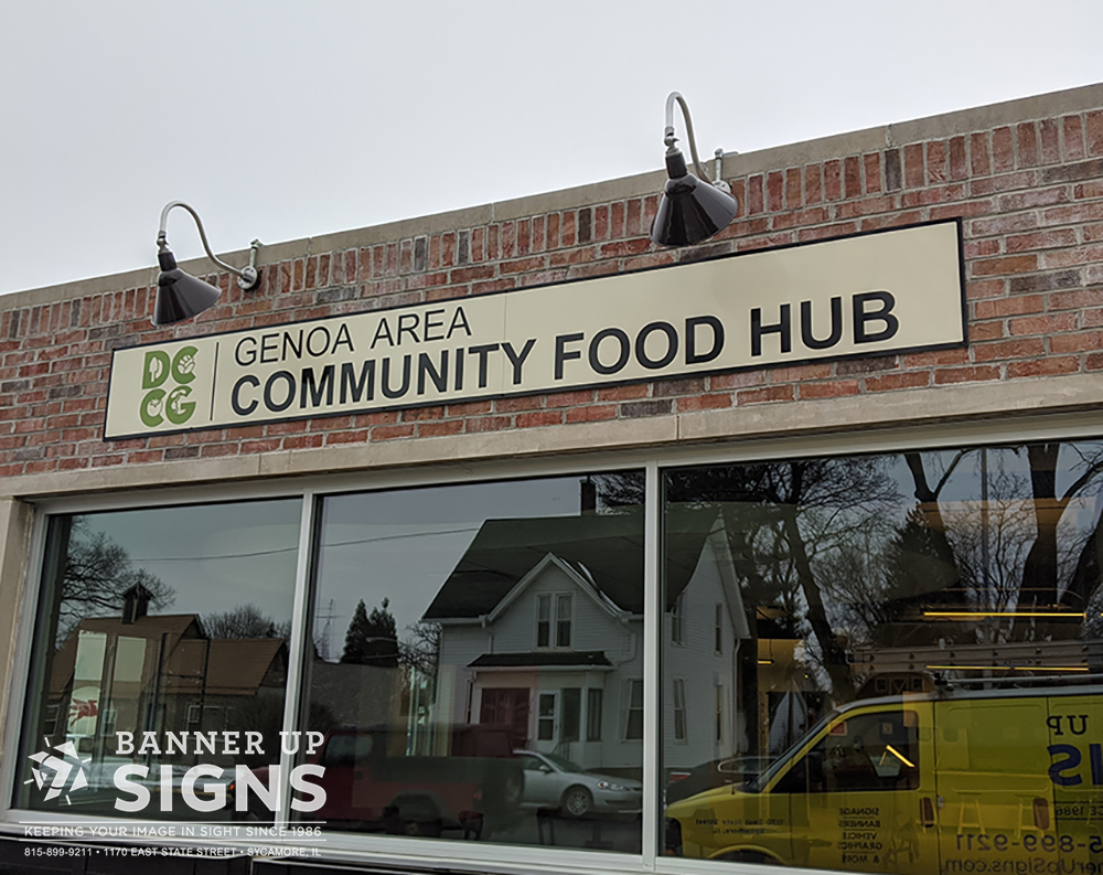 A flat aluminum sign illuminated by overhead lights informs people of the location for Genoa Area Community Food Hub.