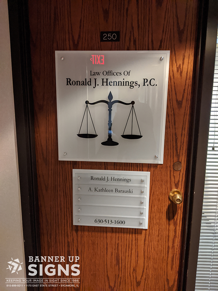 Mounted to a wood office door, a white acrylic backer with clear acrylic mounted by stand-offs has a black law logo. Below another white acrylic backer with small acrylic panels has black text of the names of the lawyers at the company.
